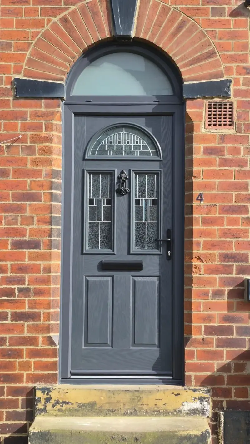 An anthracite composite door with two arched leaded panels and an ornate knocker, beneath an arched anthracite fanlight, set in a red brick arch with stone springers. The numeral 4 is fixed to the brickwork.