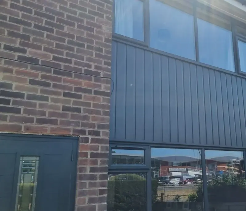 A two-storey elevation clad in vertical anthracite panels, with large anthracite-framed windows above and below, beside red brick.
