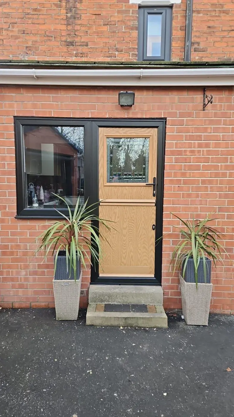 An oak-look composite stable door with a decorative glazed top panel and a black frame, beside a black casement window in red brick, with a tall wicker-effect planter either side.
