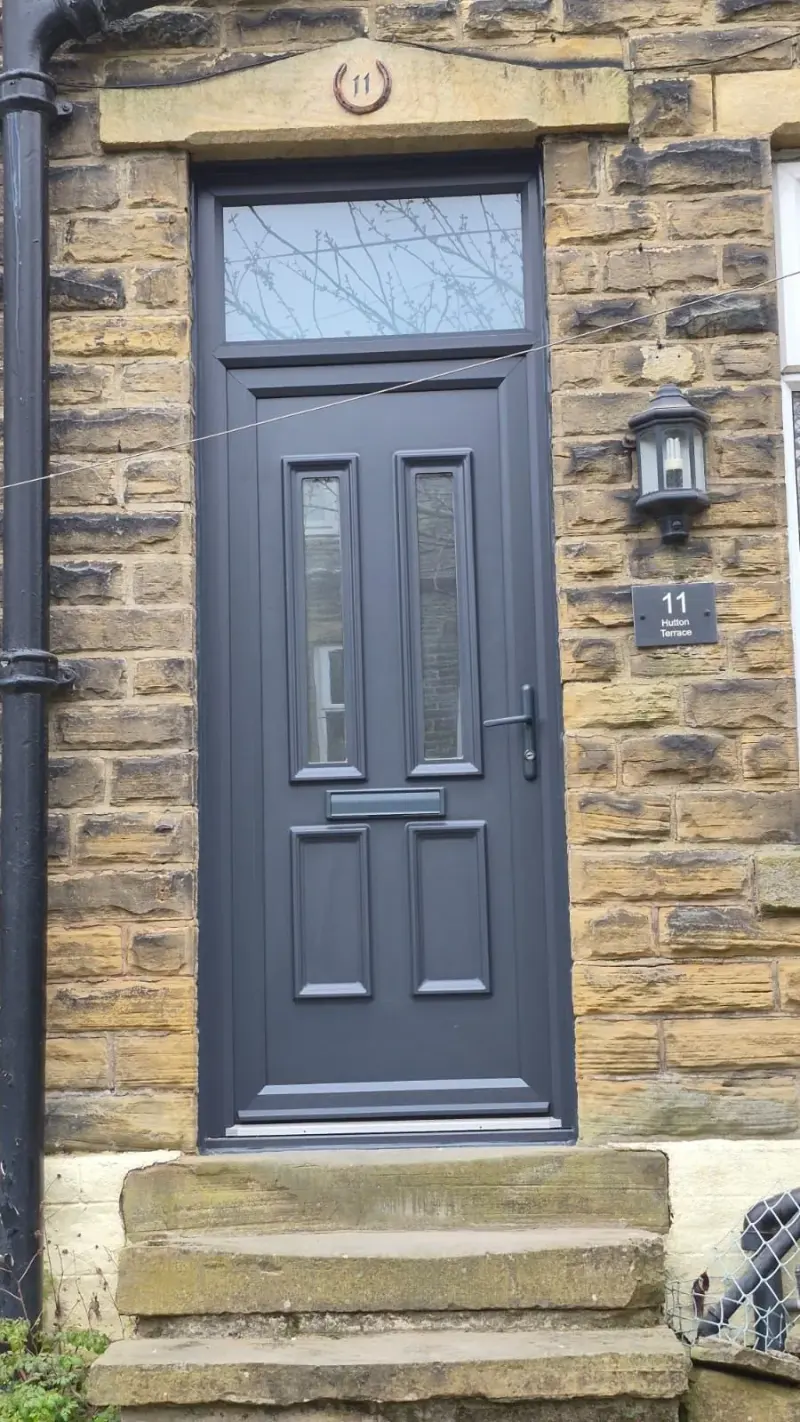 An anthracite uPVC door with two glazed panels and an anthracite transom light above, set in Yorkshire stone. A slate plaque reads 11 Hutton Terrace and a horseshoe numeral 11 sits on the lintel.