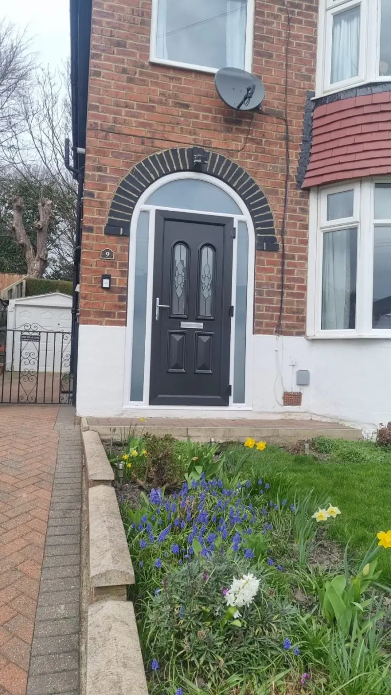 A black composite door with two arched leaded panels and narrow sidelights, set in a brick arch of alternating dark and buff voussoirs, with spring bulbs in the border.