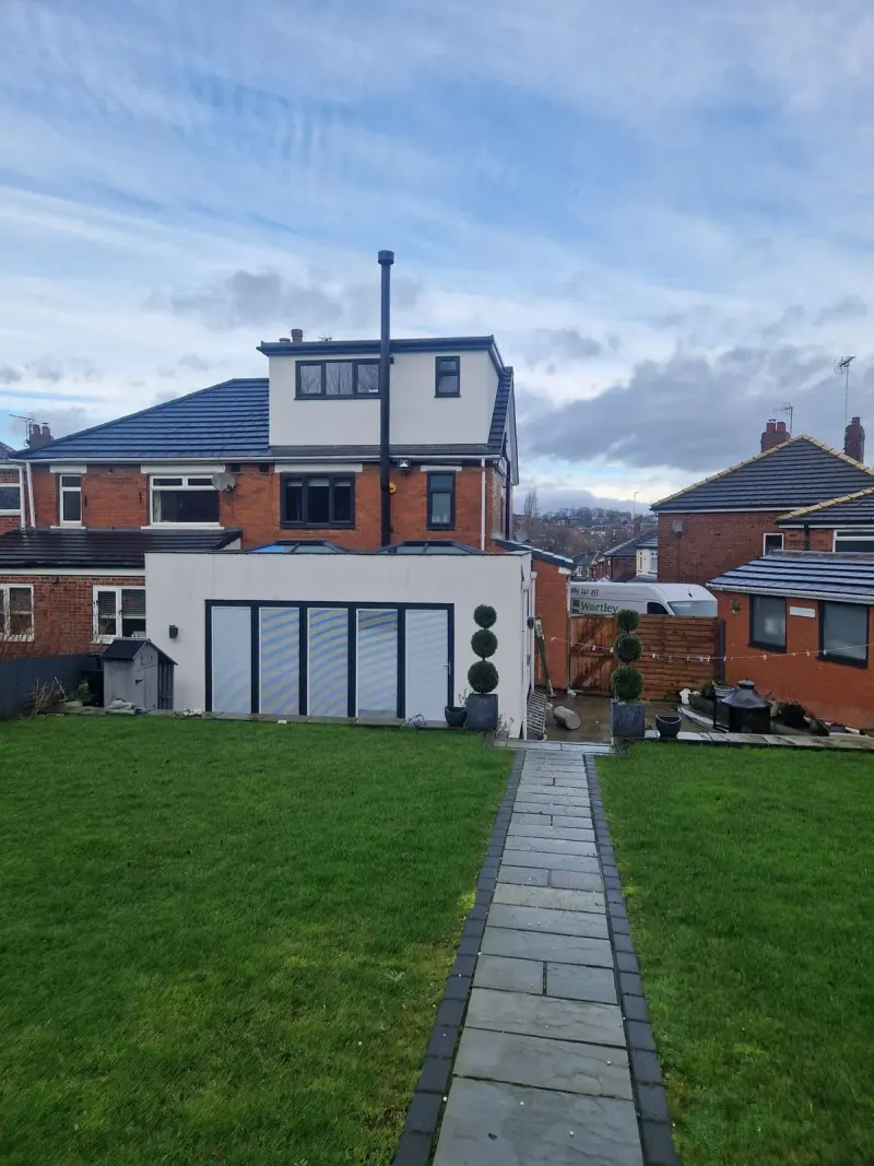 A rear garden with a paved path leading to a white-rendered flat-roof extension with dark-framed glazed doors still covered in protective film, twin roof lanterns above, and a Wortley Windows van parked beyond the fence.