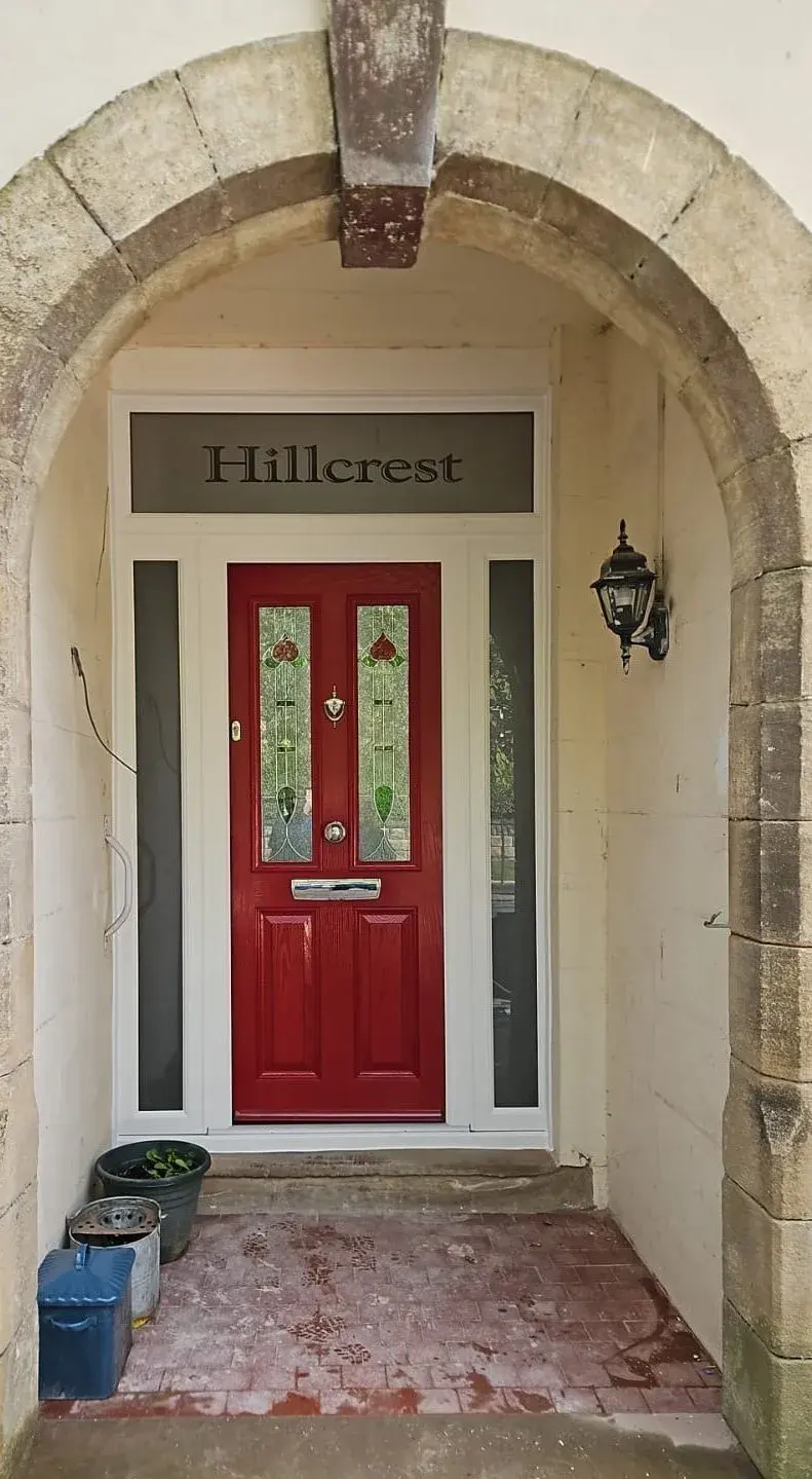 A red composite door with two Art Nouveau stained-glass panels, a brass knob and letterplate, two sidelights and an etched overlight reading Hillcrest, set in a stone arch.