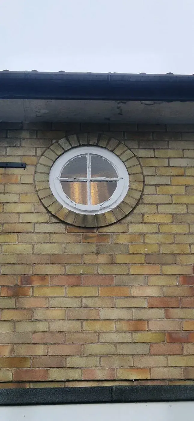 An old round porthole window with flaking paint, set in yellow brick.