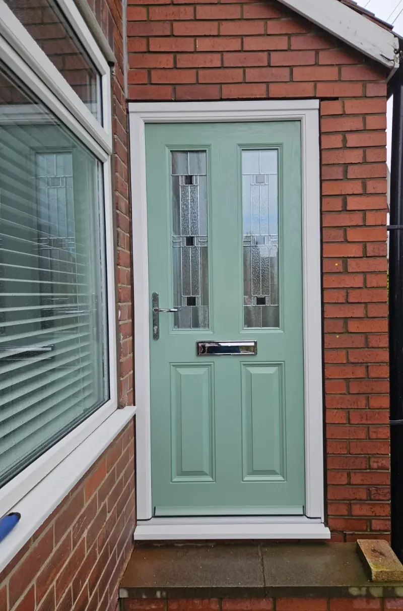A sage-green composite door with two leaded panels in a geometric pattern and chrome furniture, beside a bay window in red brick.