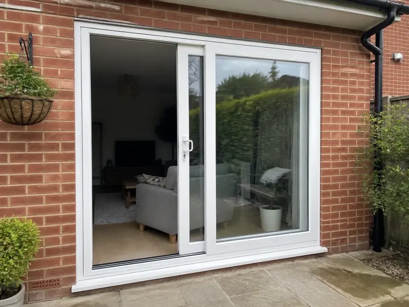 Illustrative image of a white uPVC two-panel sliding patio door partially open in a red-brick wall, looking into a sitting room from a paved garden patio.