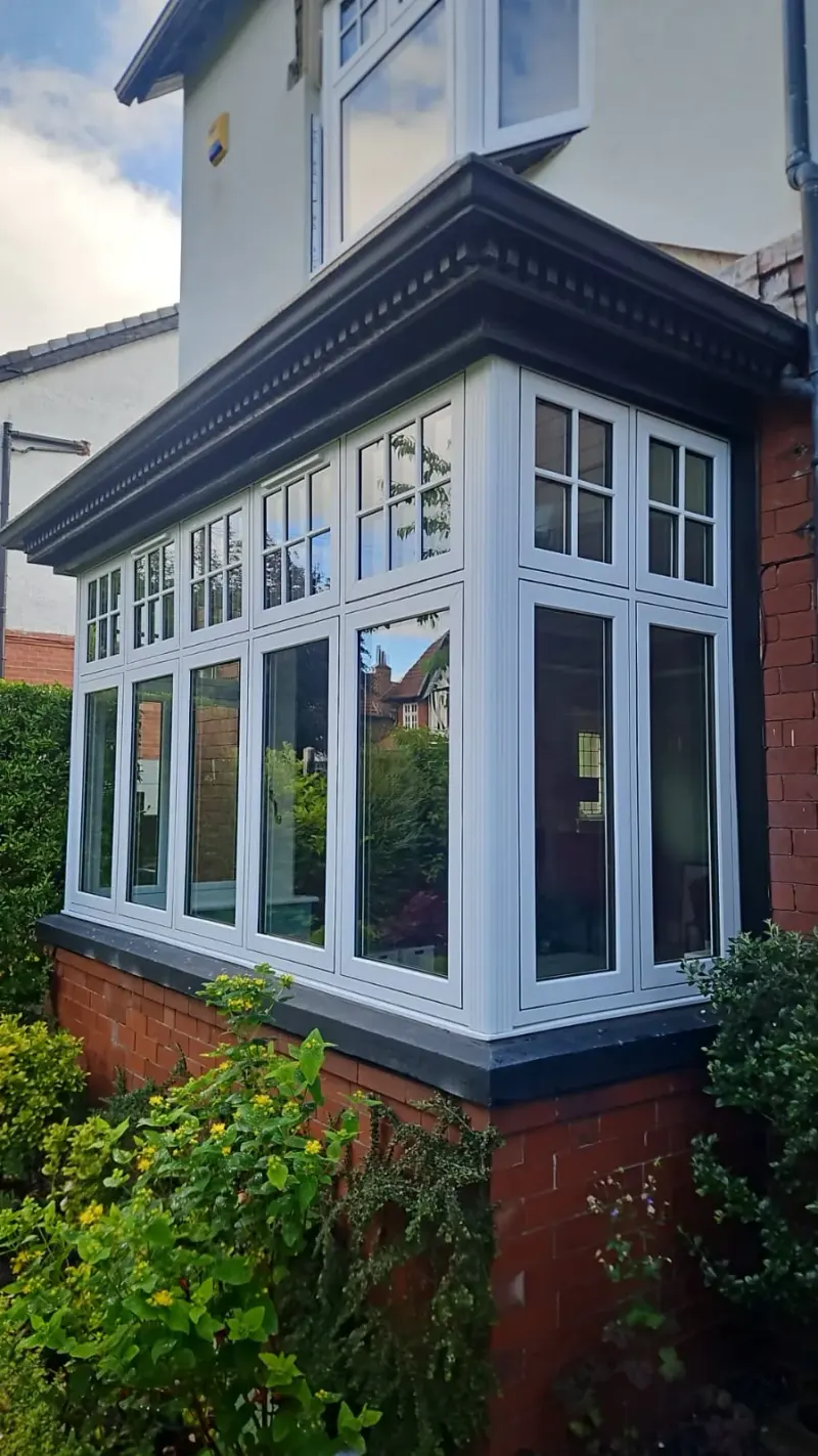 A large white square bay window with Georgian-bar top lights, projecting in flat sections from a red-brick dwarf wall beneath a dentil-moulded cornice, with shrubs in front.