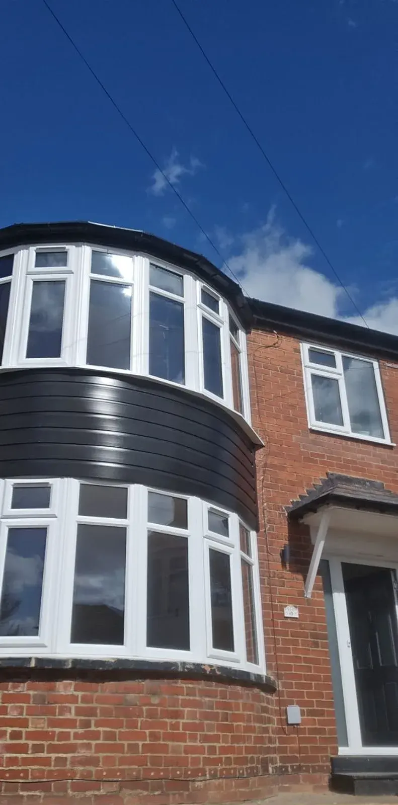 A two-storey white uPVC bow window with a black cladding band between the floors, on a red-brick semi-detached house against a clear sky.