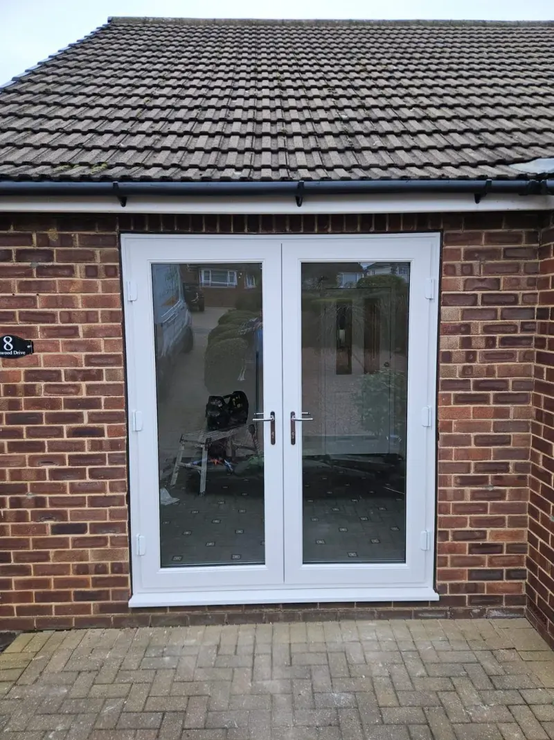 White uPVC french doors newly fitted in red brick, with a trestle and a tool bag visible through the glass and a van and the houses opposite reflected in the upper panes.