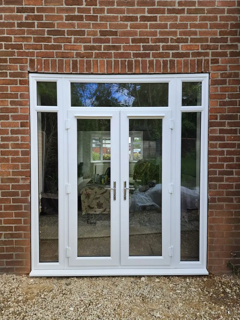 White uPVC french doors with side panels and a toplight above, set in red brick above a gravel bed.